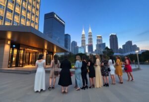 Group of women in elegant dresses pose in front of Kuala Lumpur skyscrapers at dusk.
